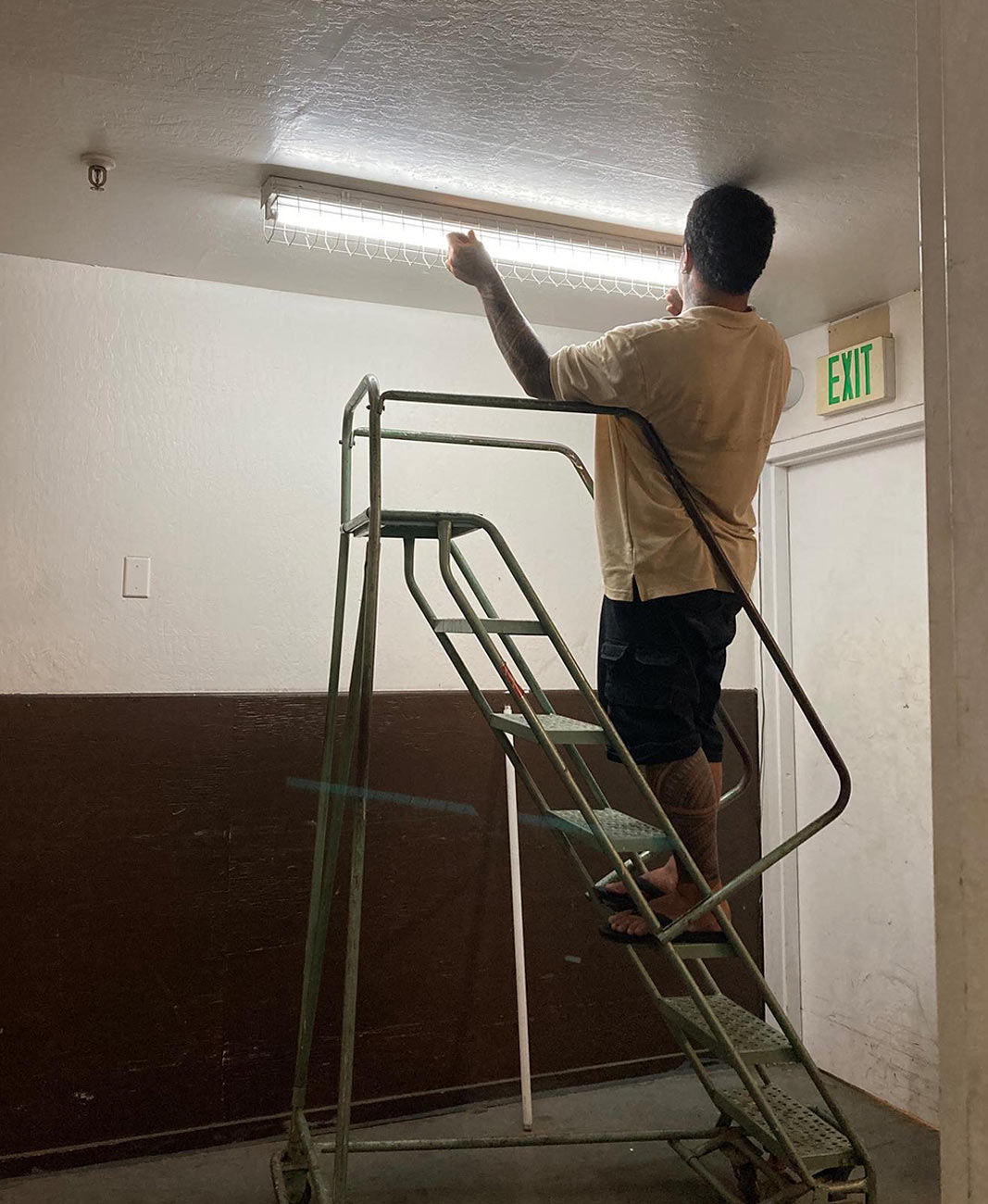 a man stands midway on a rolling ladder to change a fluorescent light in front of an exit