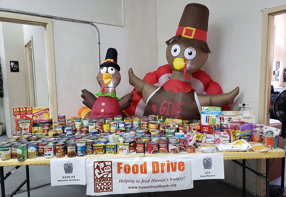 front view of a table loaded with packaged and canned food, a sign on the table reads: Hawaii Food Bank Food Drive, behind the table sit two large inflatable turkeys dressed as pilgrims