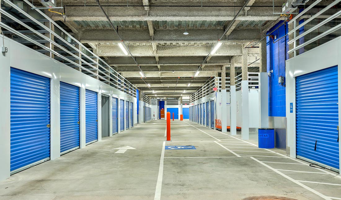 view down the wide walkway of a Morningstar storage block, the unit doors are painted a deep blue and the parking bollards and industrial flat carts are a vibrant orange