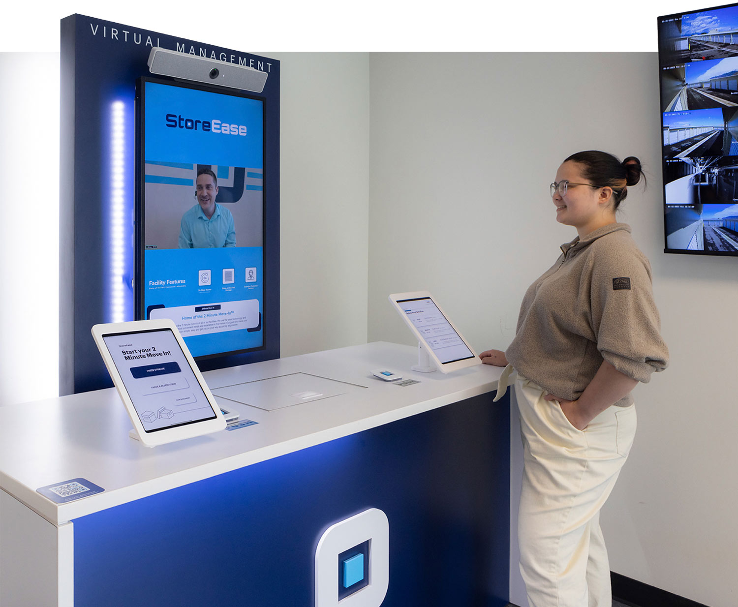 a woman smiles while standing at the StoreEase self service desk that holds two check-out tablets, behind the desk is a screen labeled Virtual Management, telecasting a StoreEase representative