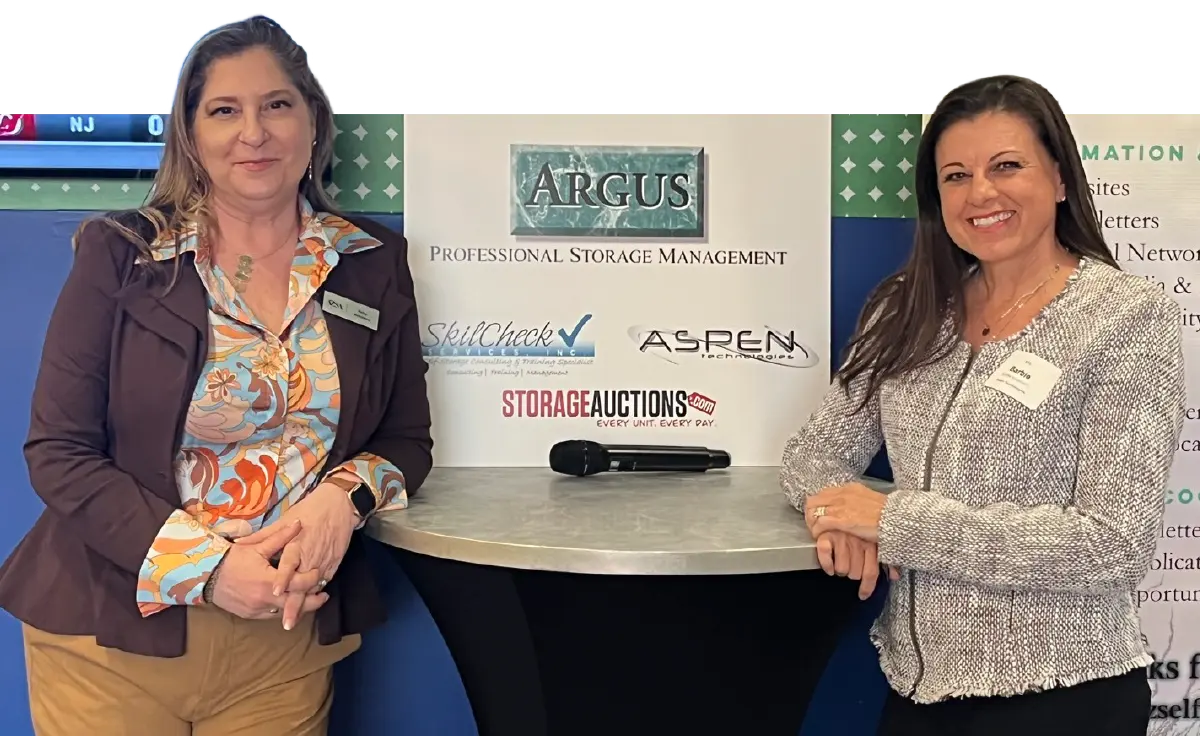 Amy Amideo smiling beside opposite another a woman as they both smile for a picture together in front of an Arguse Self-Storage Advisors Professional Storage management booth logo area