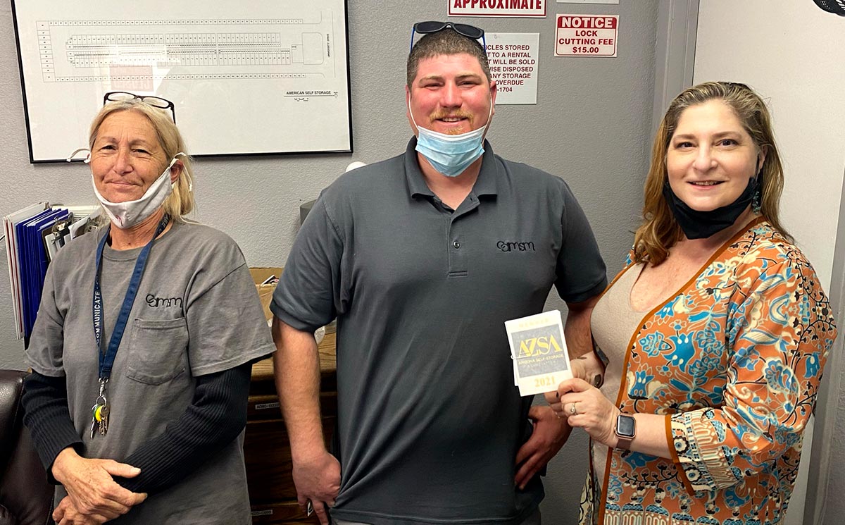 Amy Amideo smiling holding a 2021 Arizona Self Storage Association logo branded member sticker beside opposite another a woman and a man as they all smile for a picture together inside somewhere in a room