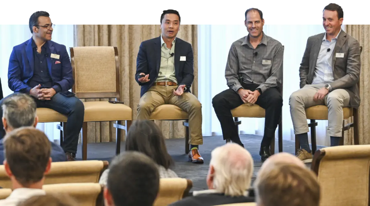 Landscape photograph of Drew Hoeven smiling (pictured to the very end far right) in a dark grey suit and light sky blue open top collar button-up dress shirt seated down in a chair next to other fellow professional work peers as part of a seminar talk meeting indoors somewhere as they glance at the audience