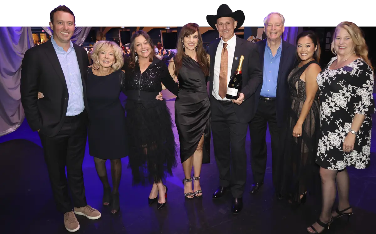 Landscape group photograph of Drew Hoeven smiling (pictured to the very end far left) in a dark black suit and light sky blue open top collar button-up dress shirt posing next to Marianne Nahin, Angie Guerin, Julie Alai, Todd Perry, Robert Bruning, Jenny Smyth, Pam Domingue as all of them are happy at a banquet ceremony indoors somewhere