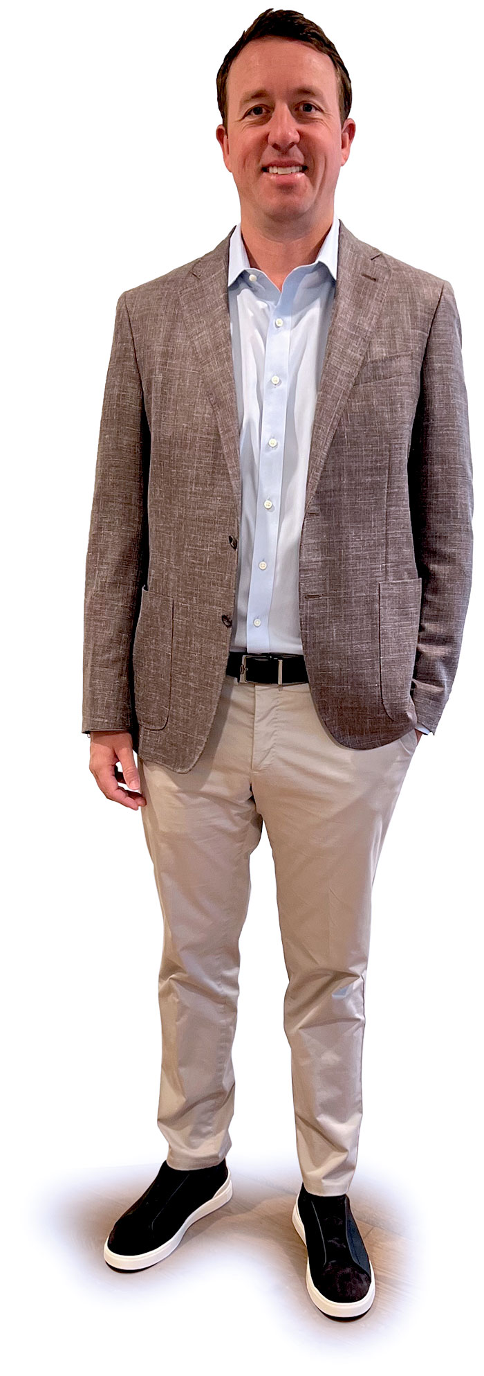 Portrait photograph of Drew Hoeven standing and smiling in a faded dark grey business blazer suit open coat and light faded sky blue button-up dress shirt as he has his left hand casually resting in his left jean pocket; he is also wearing light tan khaki jeans and black/white colored dress shoes