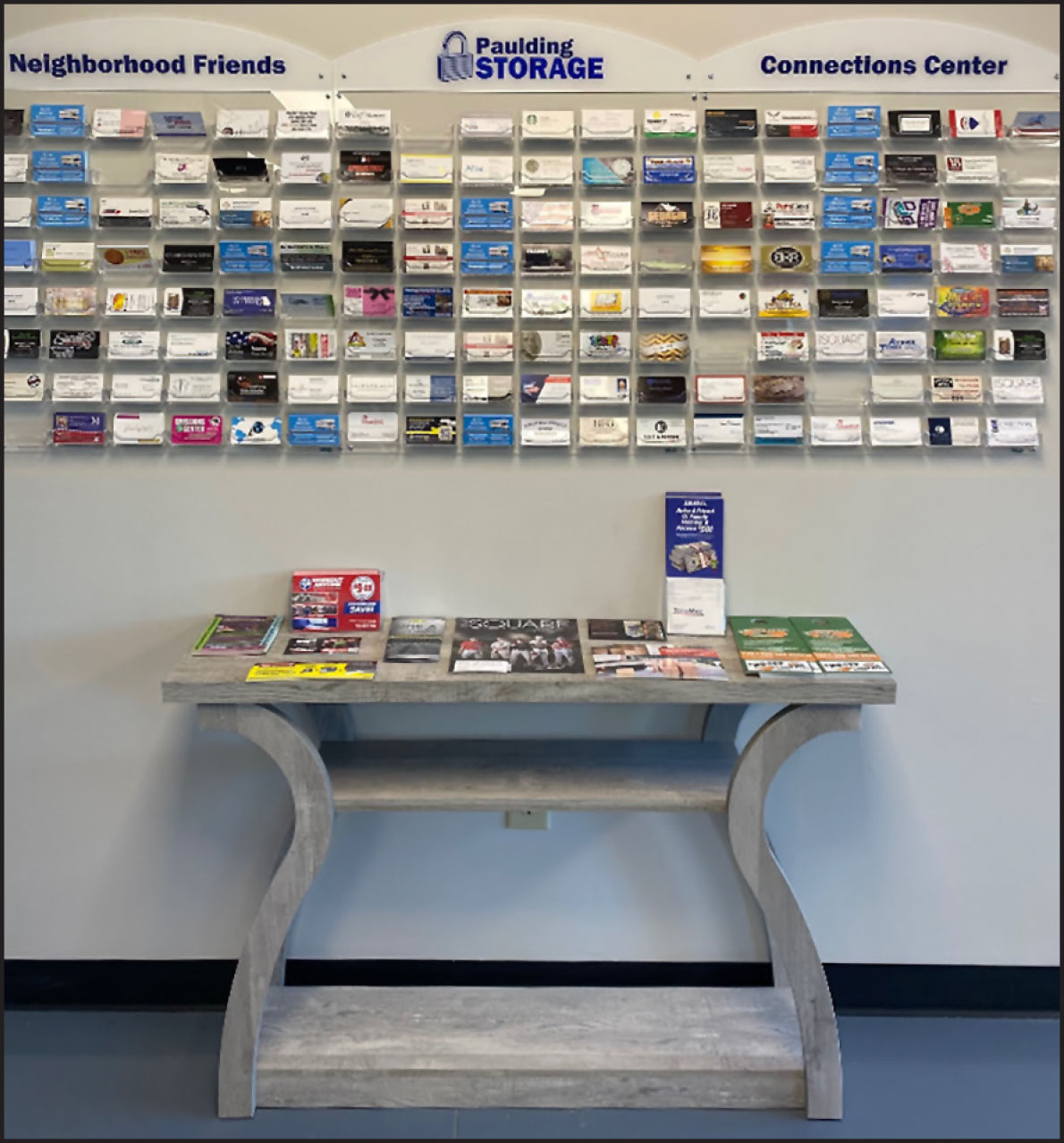 Small wooden grey table with various flyers and brochures sitting on it and shelf holding different businness cards above it
