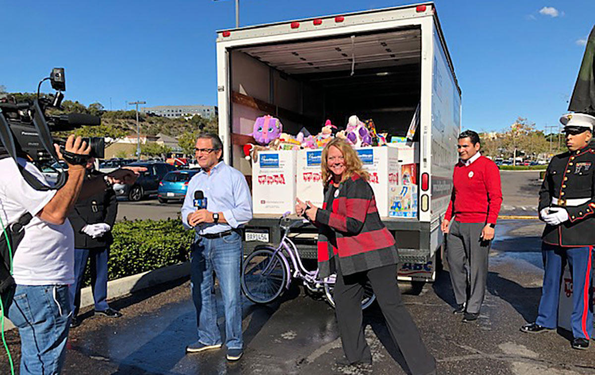 Sue giving a thumbs up next to a news reporter while standing in front of a toy for tots truck filled with supplies and toys