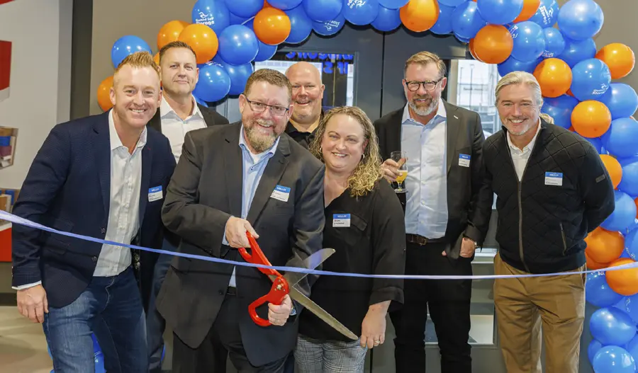 six men and a woman stand behind a blue ribbon with an arch of balloon near by, one of the men holds a large pair of scissors motioning to cut the ribbon