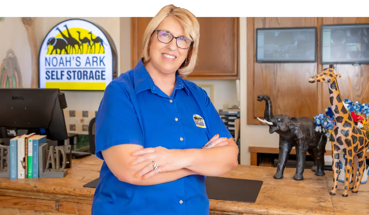 Landscape indoor photograph close-up view of Ann Parham smiling in a blue colored button-up top company polo shirt that shows the Noah's Ark Self Storage logo brand on it as she poses in front of a office receptionist desk area that has elephant and giraffe figurine statues, a computer workspace, and small books on top of a desk at a Noah's Ark Self Storage facility location