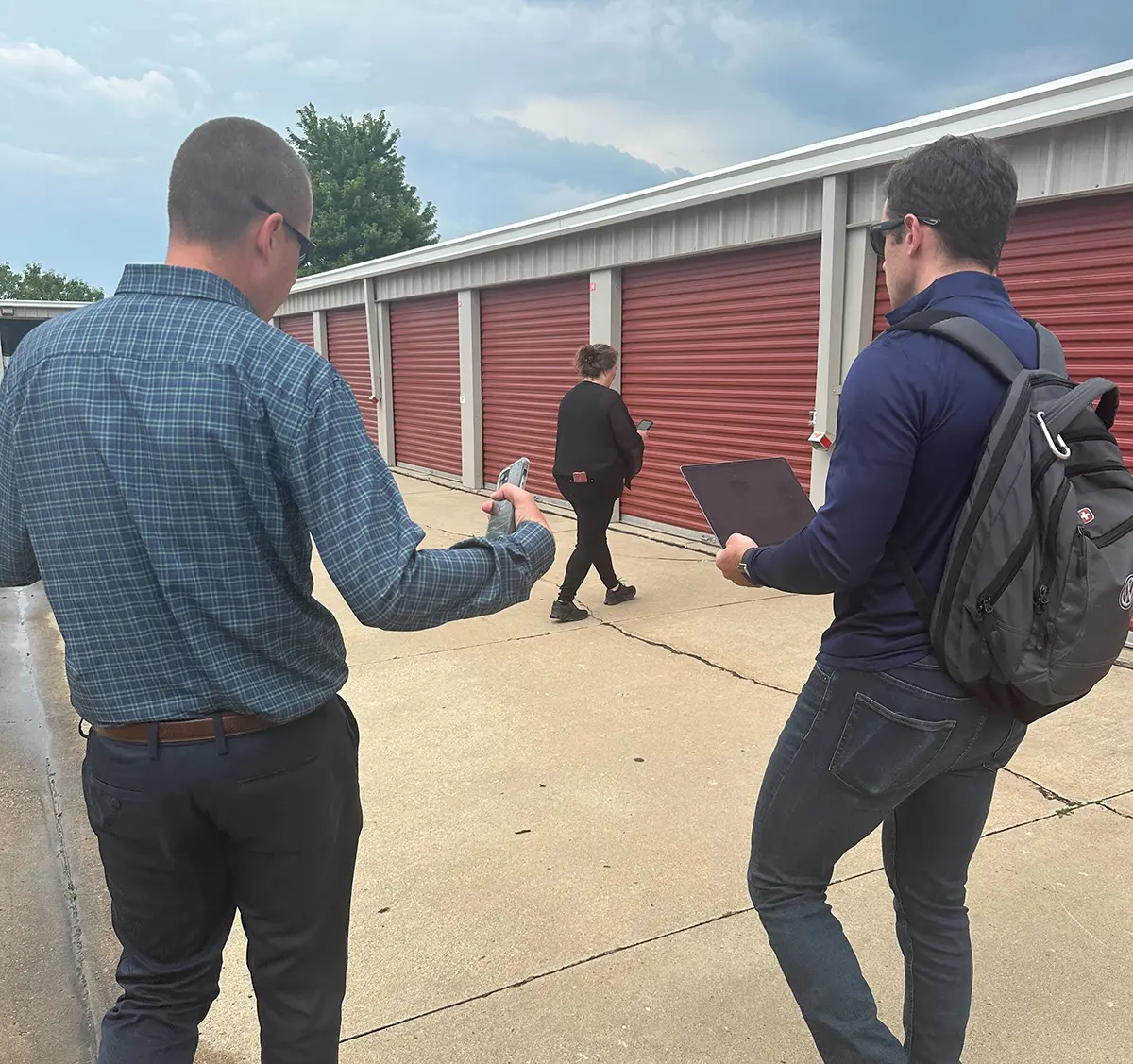 Portrait outdoor close-up photograph view of two men walking nearby towards burnt dark red self-storage units as one of these men has on black sunglasses, a dark marine blue plaid button-up dress shirt, dark navy blue chino pants, and a smartphone device of some kind in his right hand while the other man has on black sunglasses, a dark navy blue jacket, a watch on his left wrist, a dark grey/black Swissgear branded backpack, dark grey pants, and a laptop in his hands as they are glancing towards at a woman in a black blouse, black pants, and black walking fitness shoes as she has a smartphone in her hands as she is walking towards one of the burnt dark red self-storage units on an overcast gloomy day