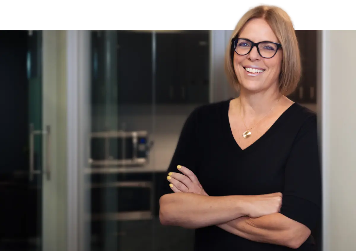 Portrait photograph of Maureen A. Lee smiling in a black v-neck blouse top, black outer frame/chrome-tinted inner frame prescription see through glasses, and a circular gold-colored necklace as she stands with her arms crossed nearby the ledge of an office room glass window