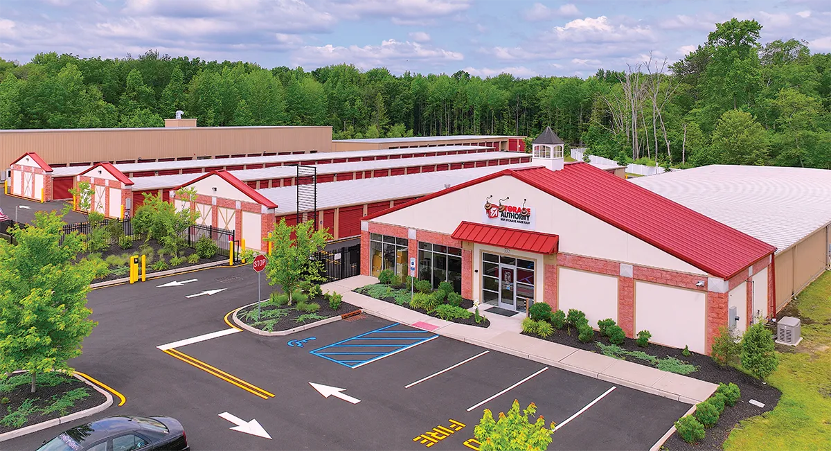 Aerial view of a self-storage facility surrounded by lush green trees. The facility has a large building with a red roof and white walls, featuring a prominent entrance labeled 'Storage Authority' with a red logo. Multiple storage units with red doors are visible behind the main building, arranged in rows. The facility is well-maintained, with landscaped greenery and a paved parking area in the foreground.