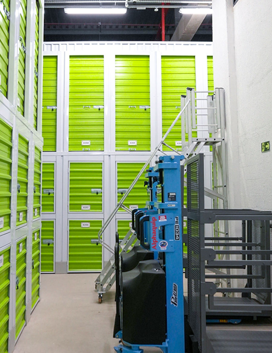 a corridor of small storage units with neon green doors in stacks of threes, a large rolling stair and a lift are parked against a wall