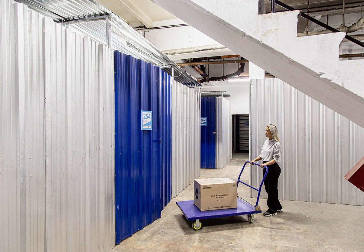 a woman in a corridor pushes a large cart holding a box towards a large blue storage unit door