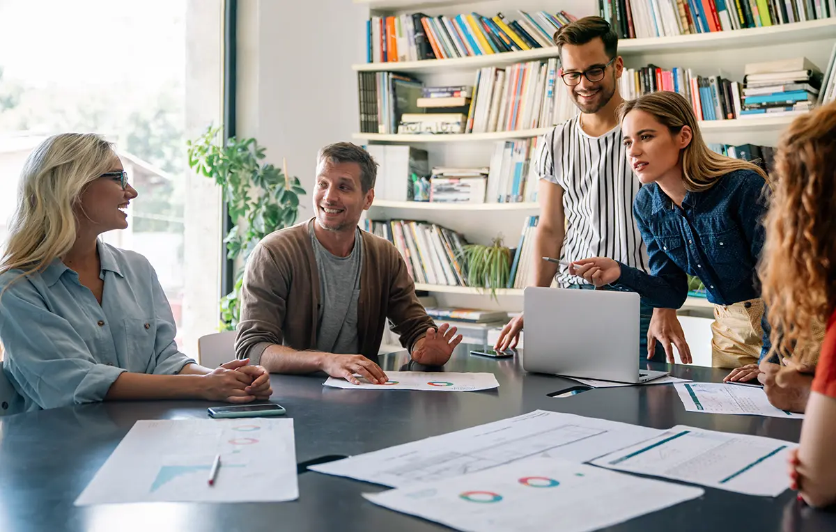 Five individuals (three women and two men) discussing about work/business at a table as there are documents of statistics/charts/diagrams nearby them while they bounce ideas off each other inside a building area of some sort