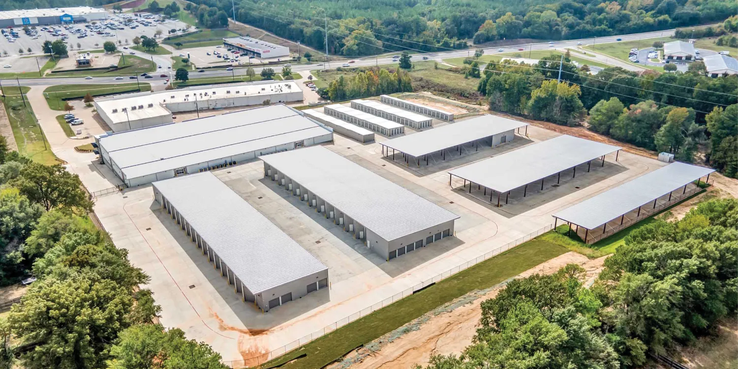 An aerial view of a large self-storage facility with multiple single-story buildings, driveways, and covered parking spaces. The facility is situated near a commercial area with trees and greenery surrounding it.