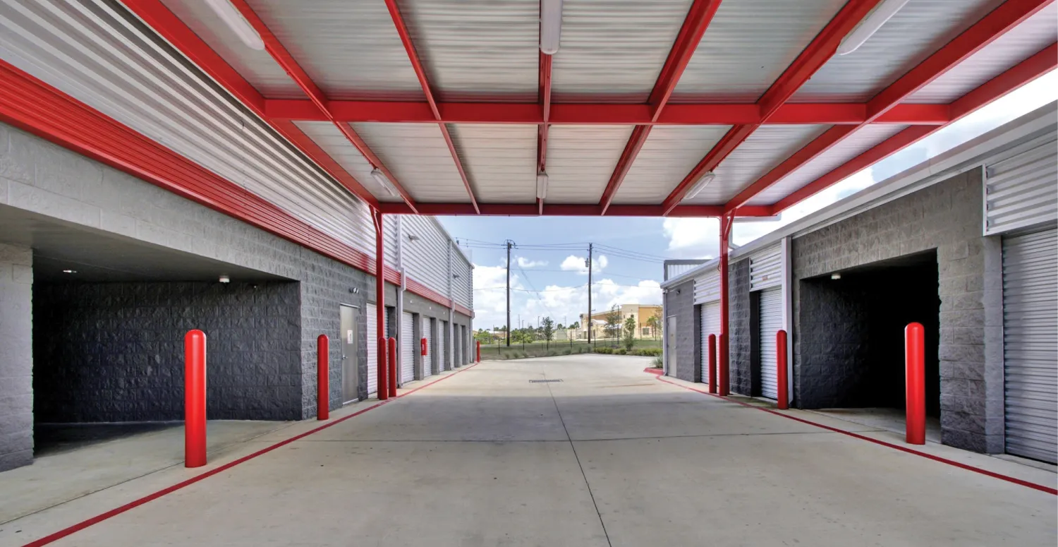 A covered drive-through area at a self-storage facility with red poles and accents, leading to storage units on either side. The walls are made of gray cinder blocks, and the ceiling is supported by red beams.