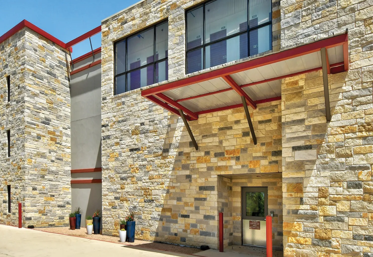 The exterior of a storage facility featuring a stone facade with red accents. The building has large windows on the second floor and a covered entrance supported by red beams.