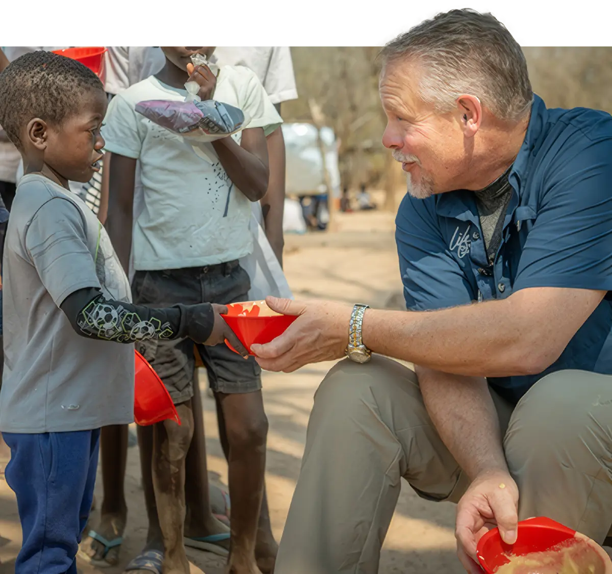 Man in a dark navy blue top and dark beige colored pants kneeling down on the dirt ground helping a small African kid by offering him a bowl of food as they look at each other