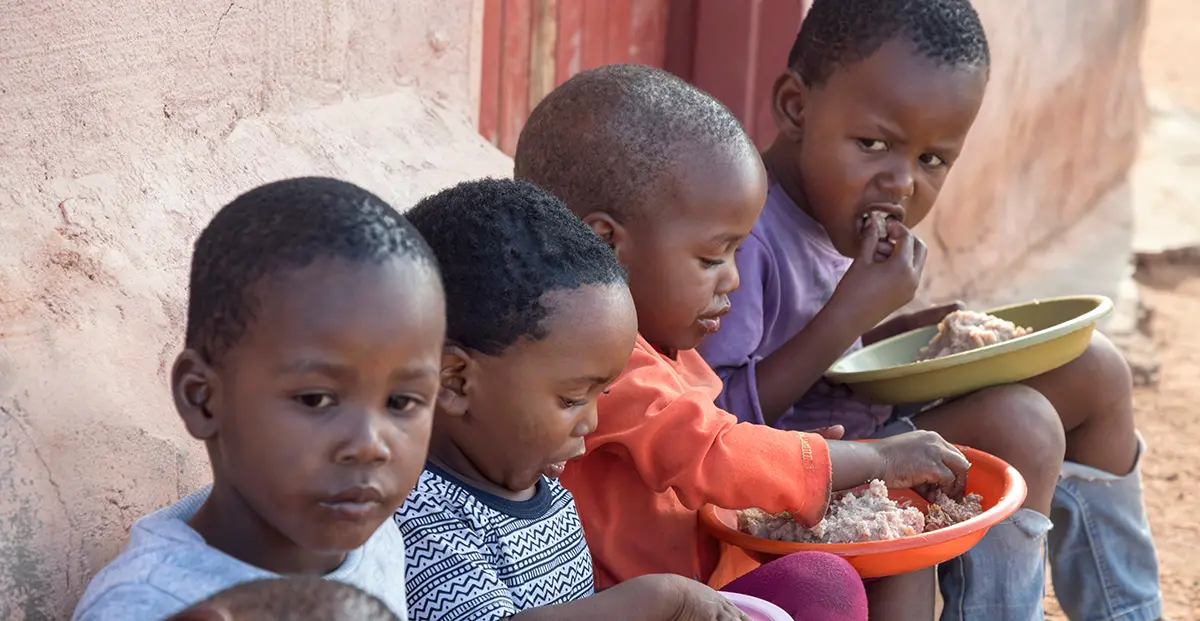 Four small African kids enjoying their food in a plate as they are seated on the dirt ground up against a wall of a building