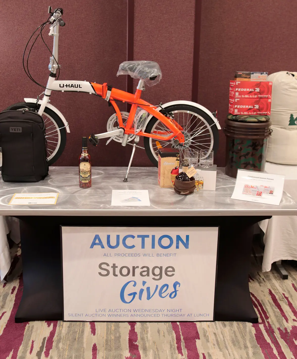 StorageGives auction items displayed on a table such as a U-HAUL branded logo small bike, a YETI backpack, other various random assorted bottles/glass cups, etc.