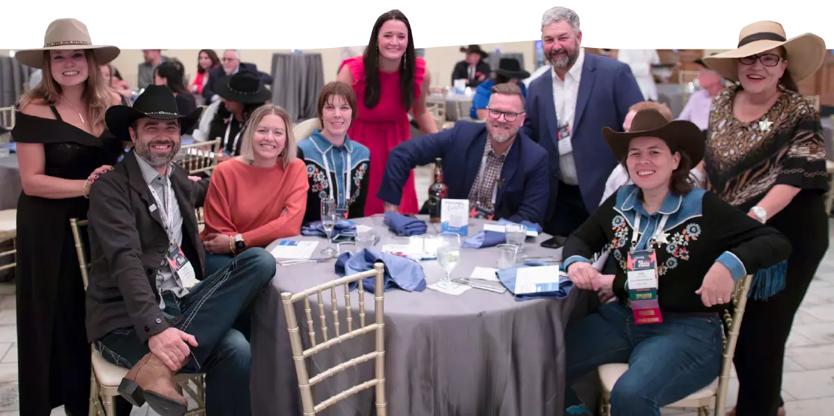 Landscape close-up photograph view of a group of nine individuals (three men and six women) smiling and posing for a picture in proper professional casual attire at a table area within a charity event of some kind held inside in a ballroom conference area