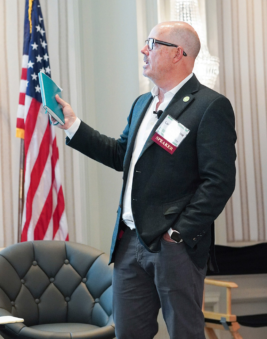 Lance speaking at conference with black suit on in front of chair and American flag