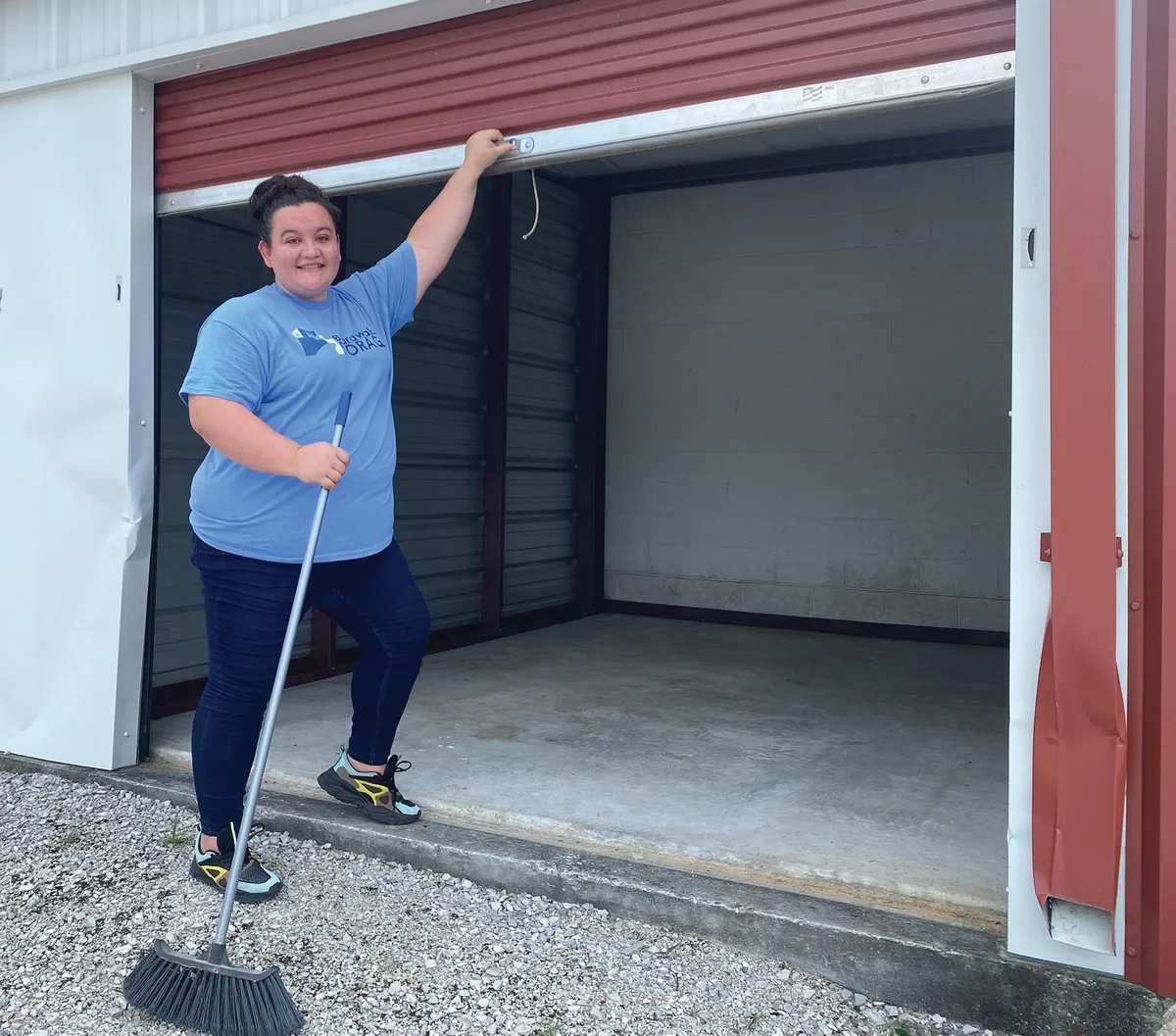 "Amy Hooks standing near her car, pointing to a rainbow in the sky. The background shows a storage facility and green hills.