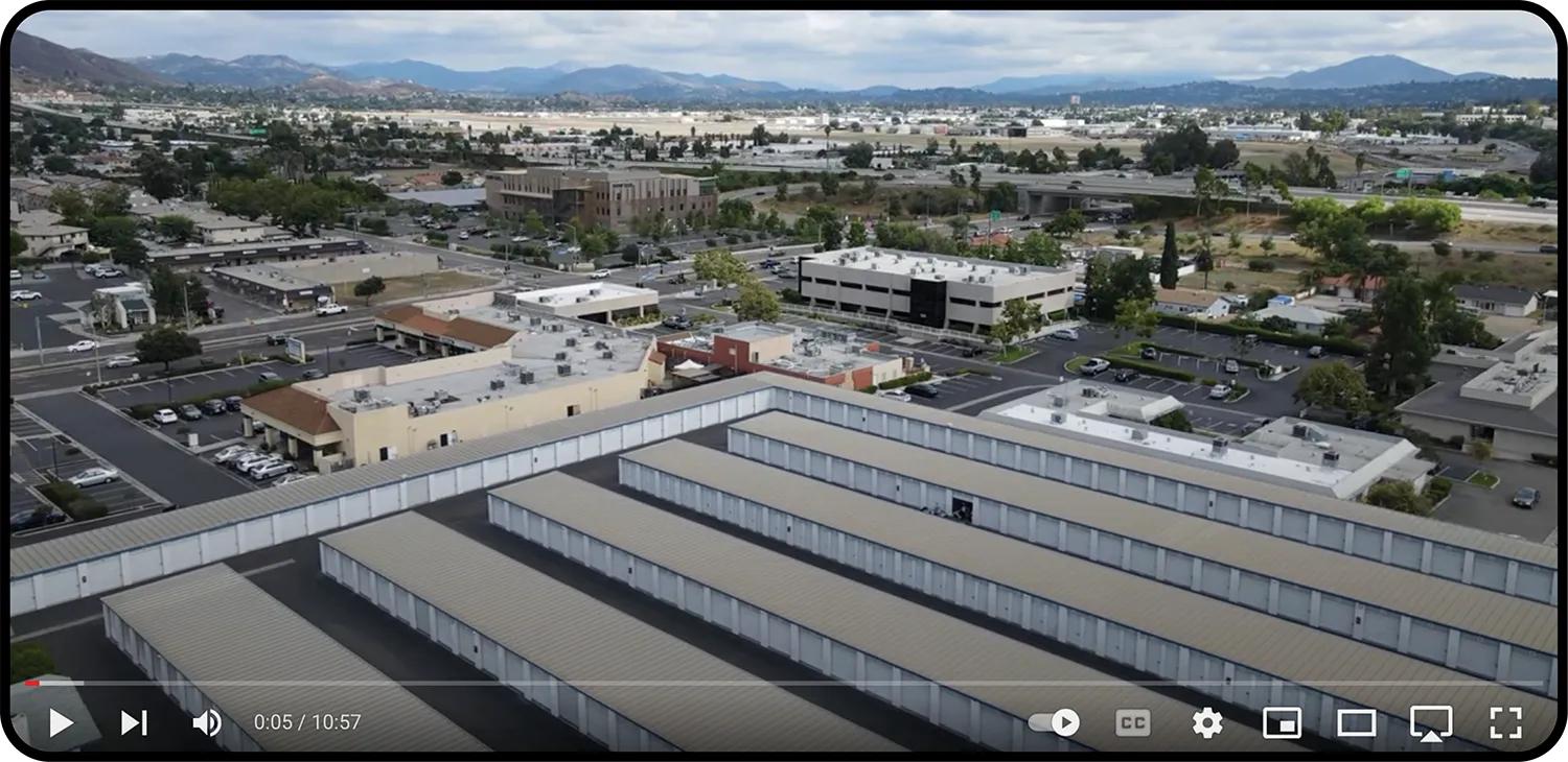 Aerial view of a commercial area with rows of storage units in the foreground. The background shows office buildings, parking lots, and a distant view of mountains under a cloudy sky. The image appears to be a screenshot from a video player with controls visible at the bottom.