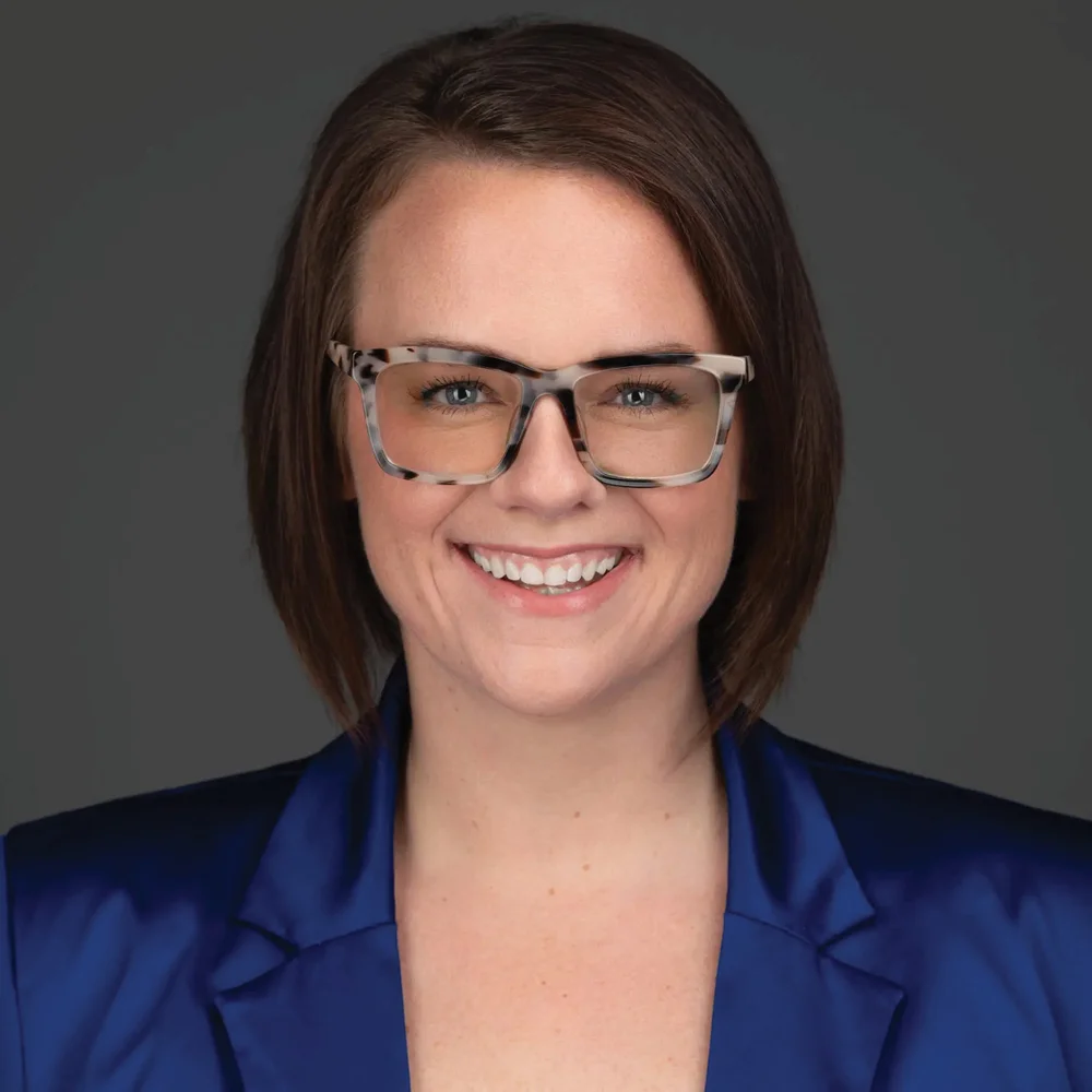 A professional headshot of a smiling woman wearing glasses and a blue blazer, set against a dark background.