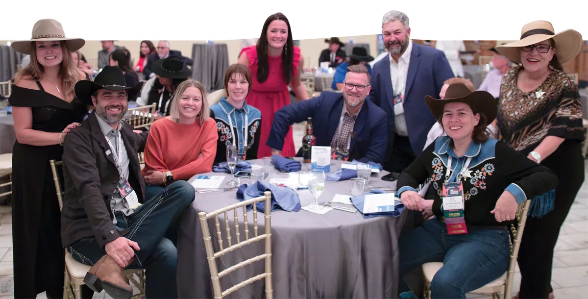 A group photo of attendees at a conference, gathered around a table. The group is smiling, with some wearing cowboy hats. The setting appears to be a formal dinner or event.