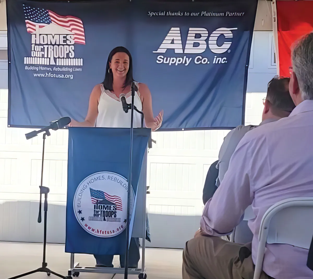 A speaker at a Homes for Our Troops event, standing behind a podium, delivering a speech. A banner in the background displays the Homes for Our Troops logo and sponsor ABC Supply Co. Inc.