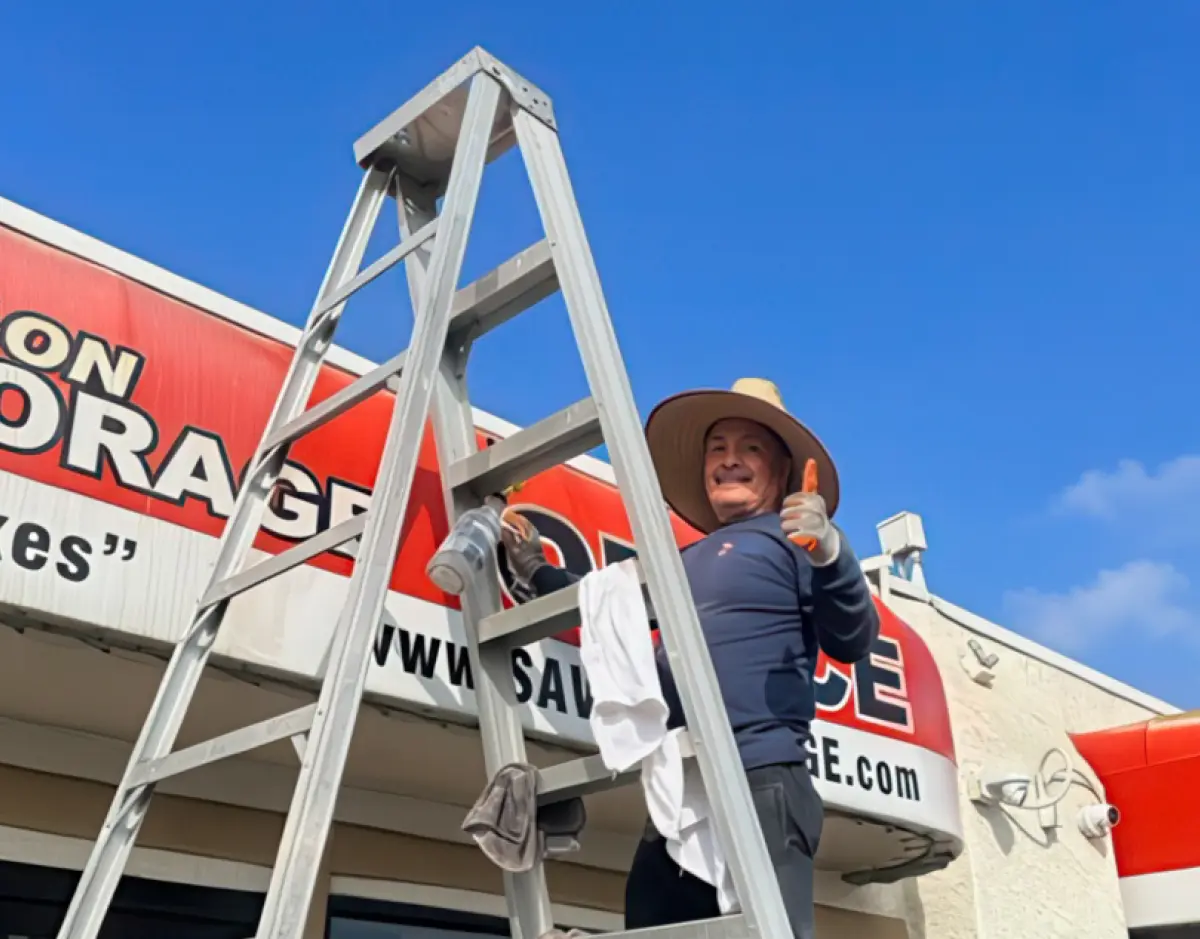 Portrait orientation exterior angle photograph outdoor view of a Sav-On Storage facility building location as right beside next to the entrance sign nearby the top of the building there happens to a be a handyman putting a thumbs-up gesture as he is standing on top of a tall ladder