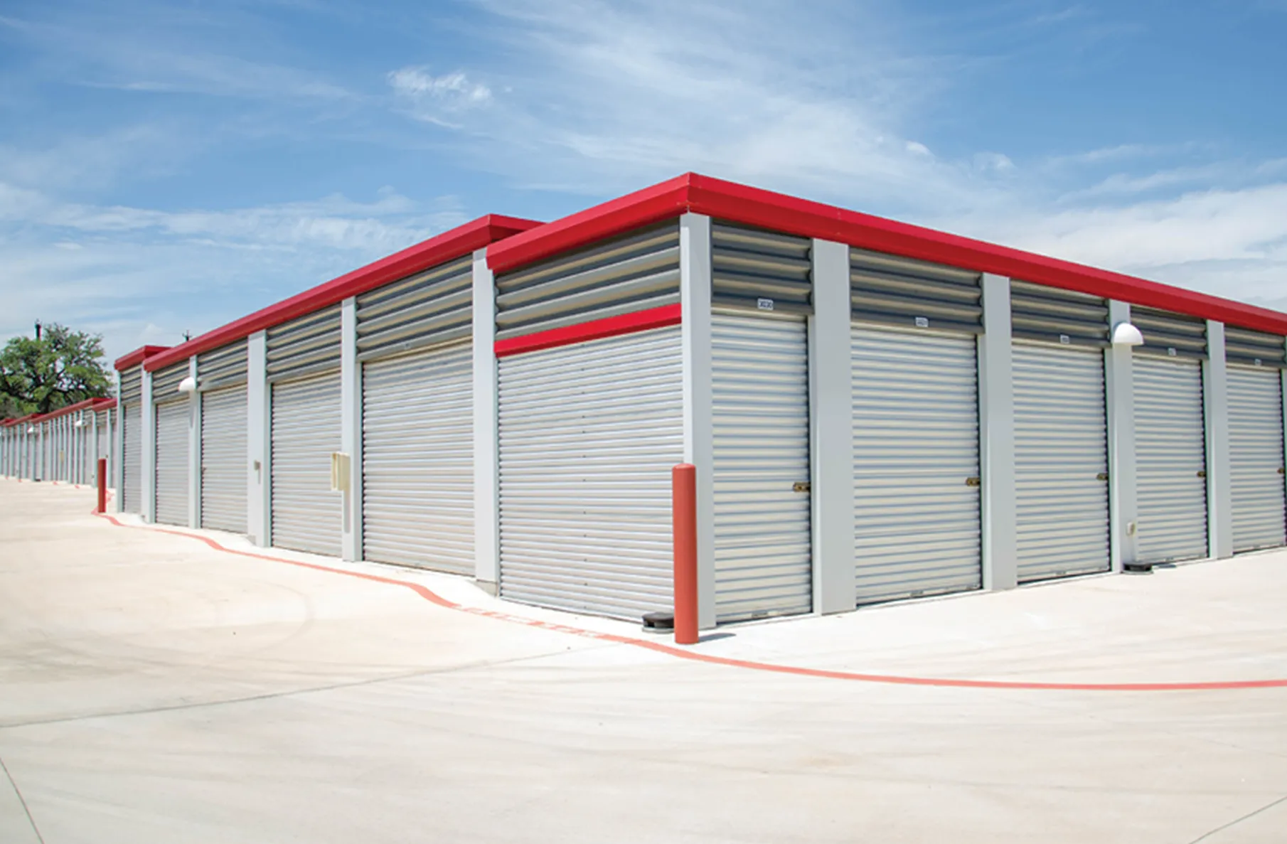 A single-story self-storage facility with roll-up metal doors and a gray exterior accented by red trim. The facility has a clean, wide concrete driveway under a bright blue sky.