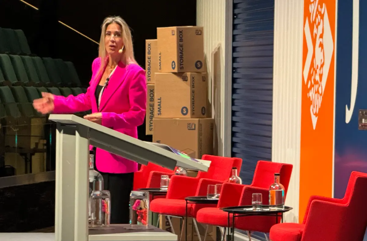 Landscape orientation indoor photograph view of Dr. Christina Radics speaking at the self-storage 2024 FEDESSA (Federation of European Self Storage Associations) conference in Stockholm, Sweden within an auditorium area as on the front stage there's Janus International Self-Storage promotional items and signage also