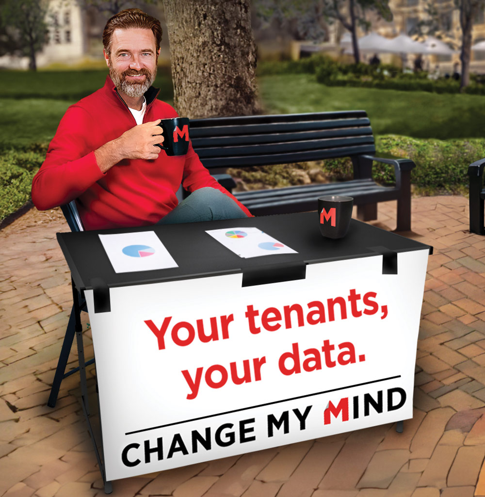 A man in a red sweater holds a mug at a table with charts and a sign in a park setting.