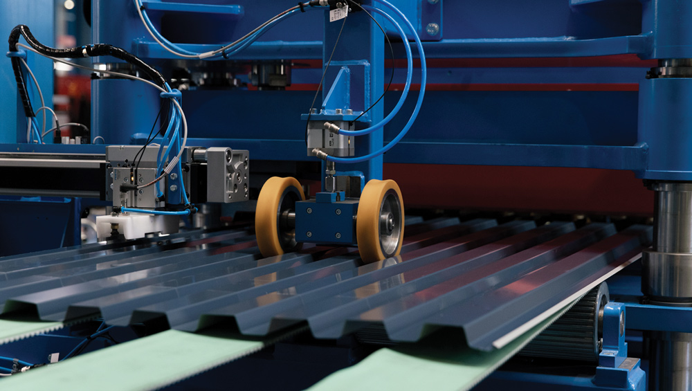Corrugated metal sheets move under robotic rollers on a blue industrial machine with wires and sensors.