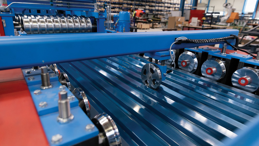 Blue corrugated metal sheets move through a blue industrial machine with multiple rollers.