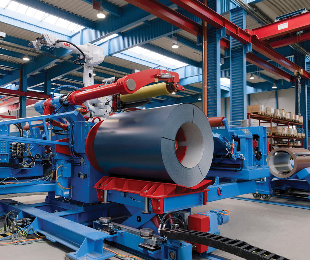 A robotic arm with a red and yellow tool manipulates a large coil of gray metal on blue machinery.