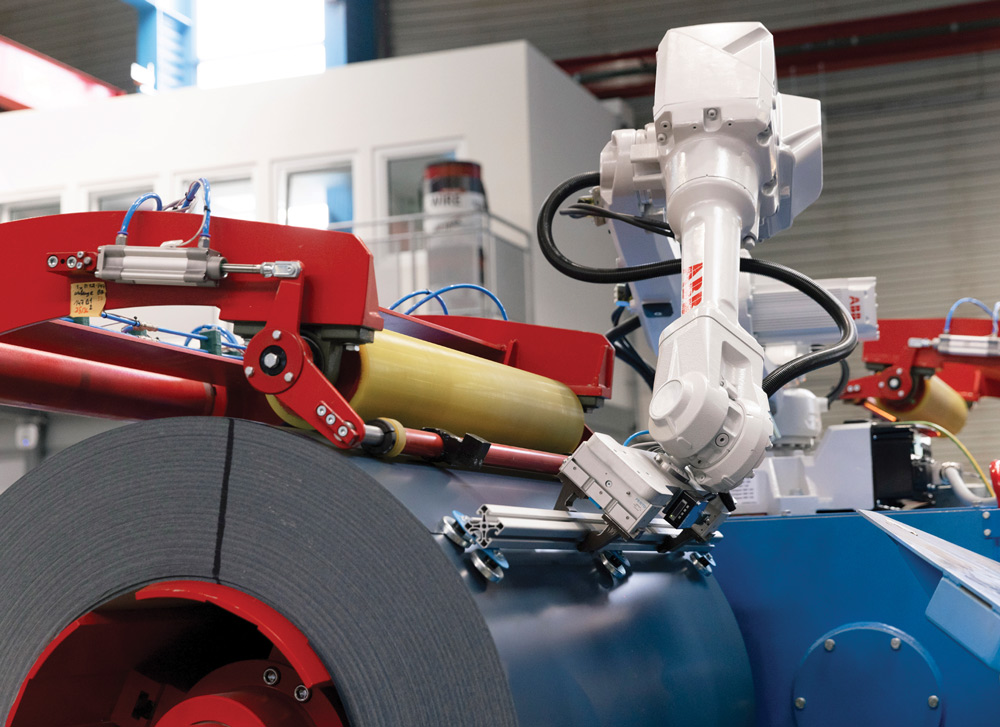 A white robotic arm with a tool interacts with a large coil of gray metal on red and blue machinery.