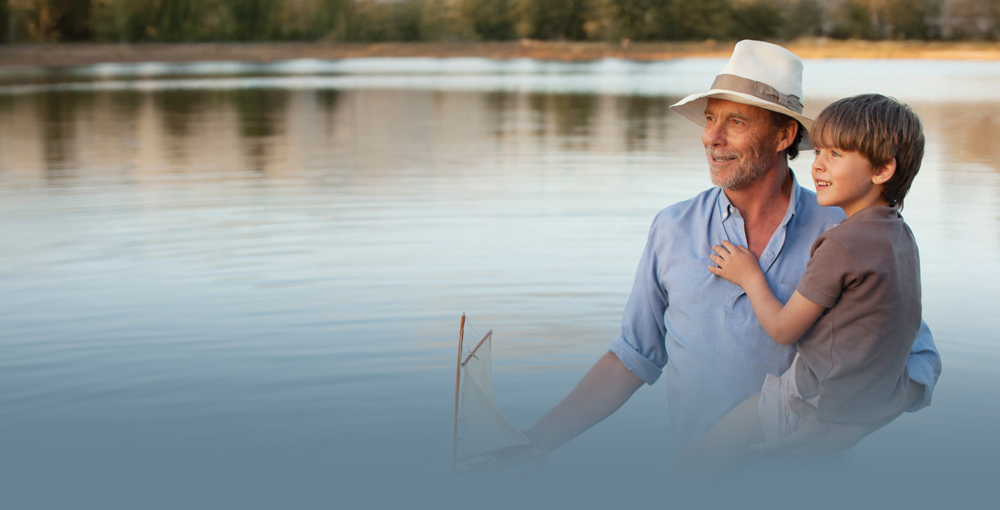 A smiling older man in a hat holds a young boy and a small sailboat near a calm lake.