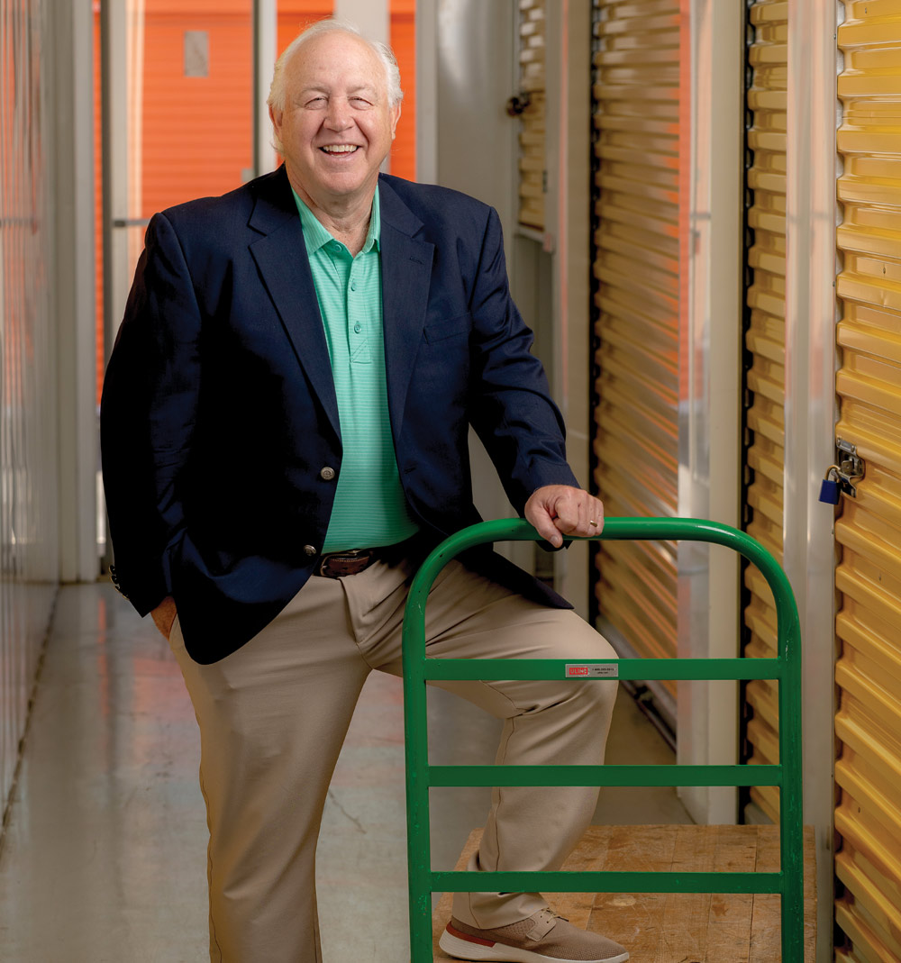 An older male in a navy blazer stands smiling next to a green hand truck, between yellow storage unit doors.