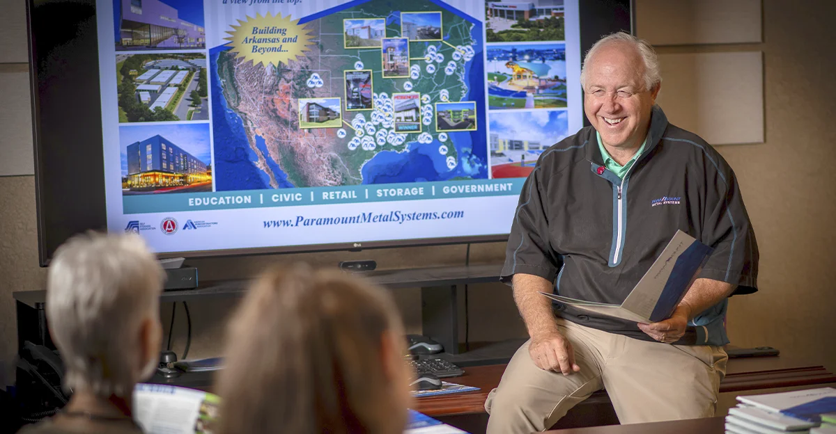 David Dodge sitting on a table and smiling while a screen shows the Paramount Metal Systems website behind him