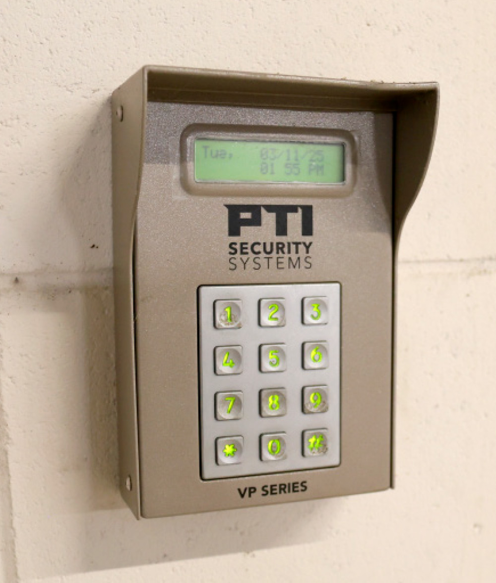 A close-up shot of a PTI Security Systems VP Series keypad with illuminated green buttons, mounted on a light-colored wall.