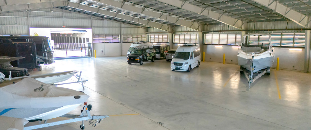 A wide interior shot of a large, high-ceilinged storage facility with a concrete floor, housing various vehicles. On the left, a large black RV and a boat covered with a white tarp are visible.