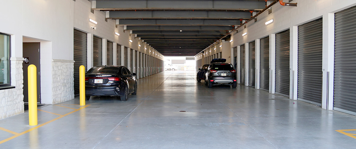 A long, brightly lit indoor corridor of a self-storage facility. On both sides, rows of grey roll-up storage unit doors are visible.