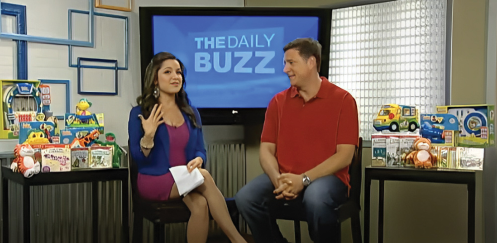 Two people sitting on a studio set of "The Daily Buzz" with toys displayed around them.