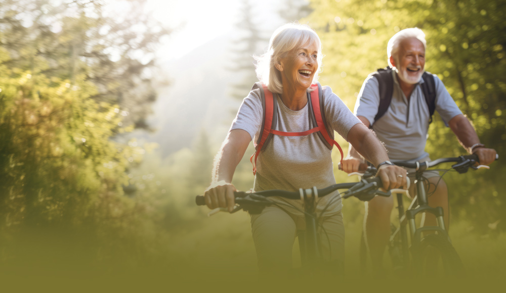 Two older adults happily cycling on a sunlit forest trail.
