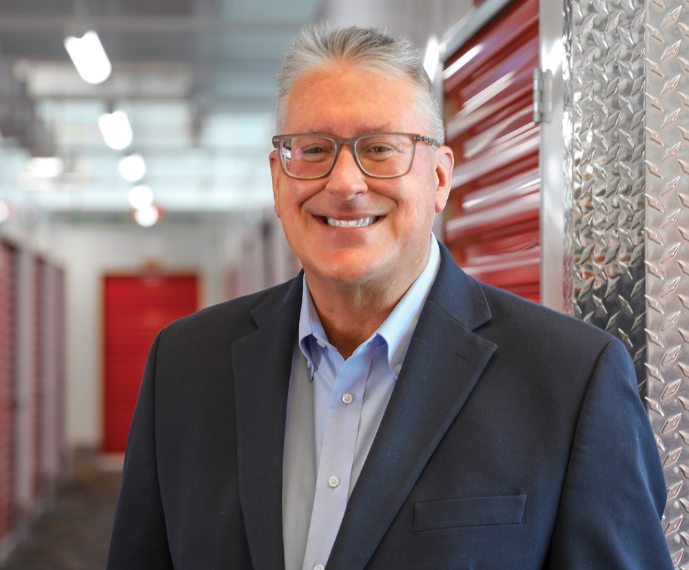 Smiling man in a suit standing in a corridor with red doors and diamond plate walls.