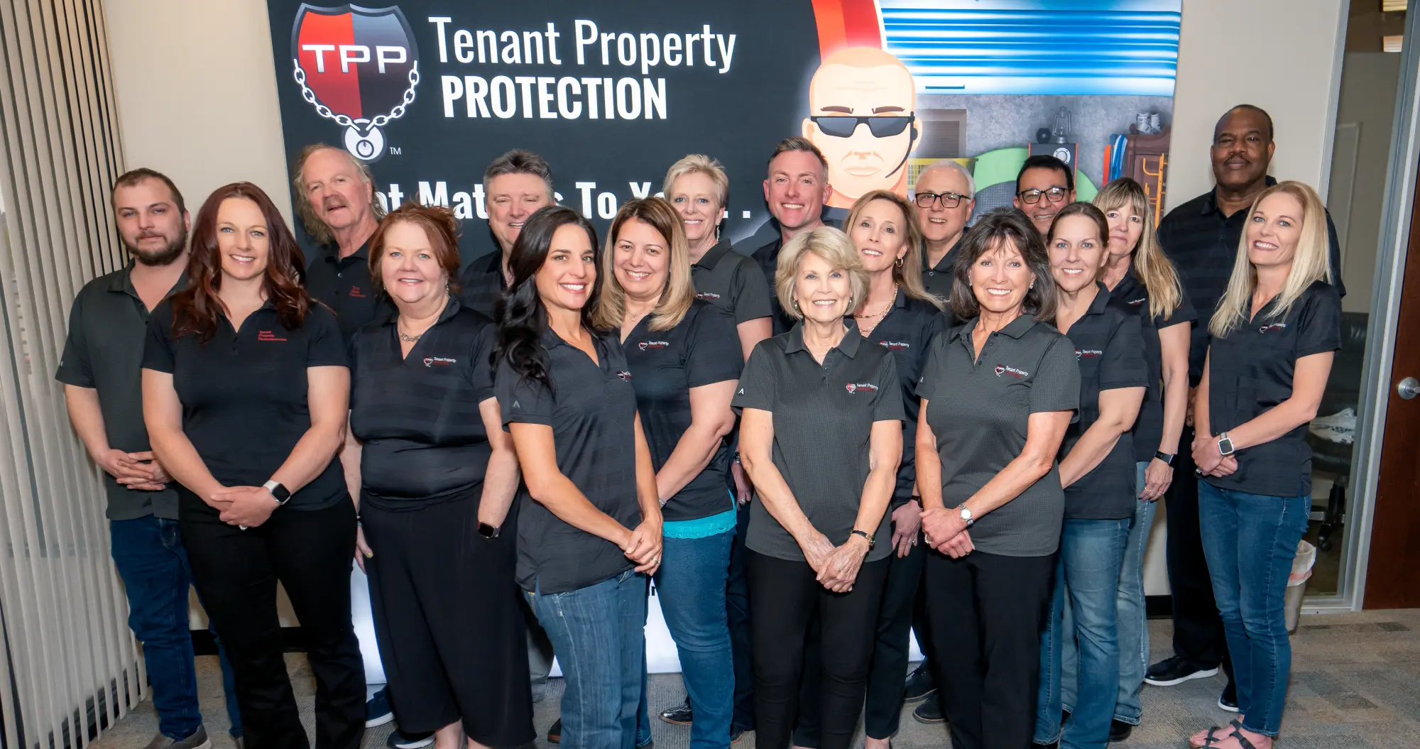 A group of 16 individuals in matching dark gray polo shirts standing in front of a banner displaying "Tenant Property Protection."